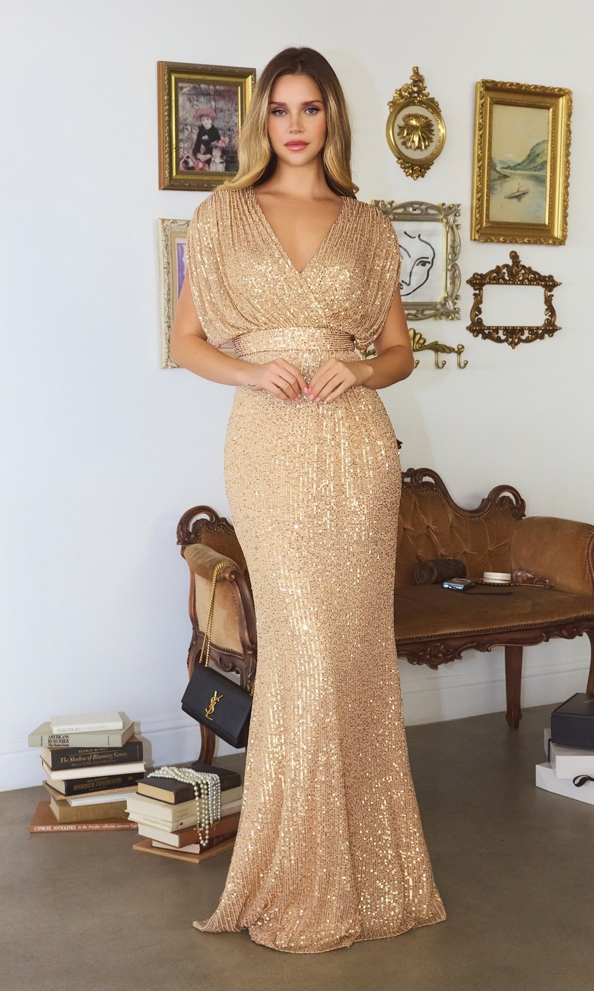 A woman wears the Cap-Sleeve Long Sequin Formal Dress: Amoris T101 in gold, standing in a stylish room with vintage decor, framed art, a brown sofa, and stacked books.